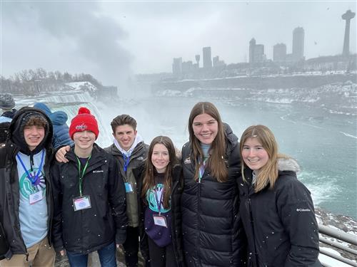 A group of teenagers in winter wear stand smiling together in front of a river covered in fog with buildings in the background. 