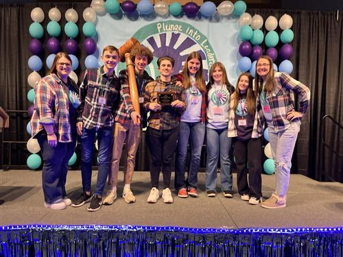A group of high school students, one holding a wooden gavel half the size of his body, stand smiling in front of a purple, blue and white backdrop that says "Plunge into Leadership."