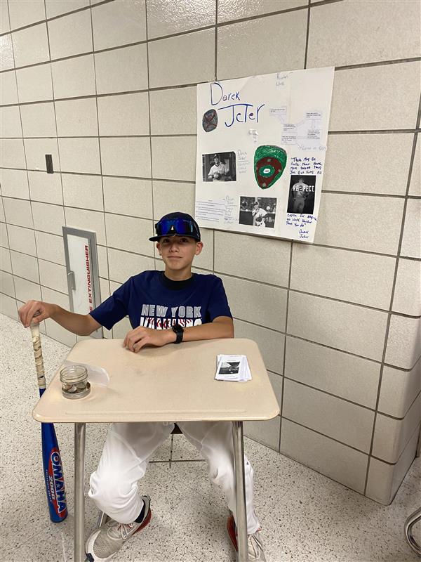 A student in a baseball uniform holds a baseball bat while sitting at a desk in a school hallway. A sign behind him reads "Derek Jeter."