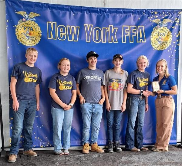 A group of high school students stand together smiling in front of a backdrop that says "New York FFA." A person presents them with an award.