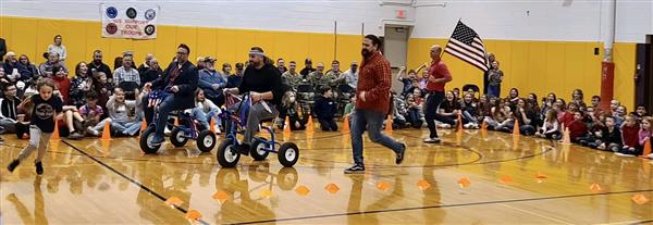 Elementary school children sit on a gymnasium floor watching adults ride tricycles between cones with an American flag in the background.