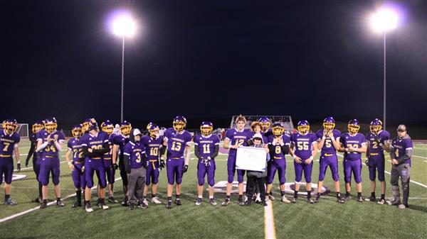 A football team in purple uniforms stand together holding a banner under bright lights in a football stadium.