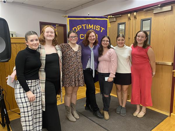 A group of high school students stand smiling in front of a purple banner that says "Optimist Club."
