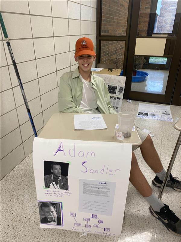 A student wears a baseball cap and a button-up shirt over a white tee shirt while sitting in a desk. A poster in front of them says "Adam Sandler."