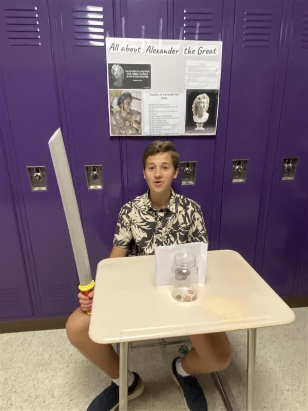 A student with a sword sits at a desk in a hallway in front of purple lockers and a sign that says "All about Alexander the Great."