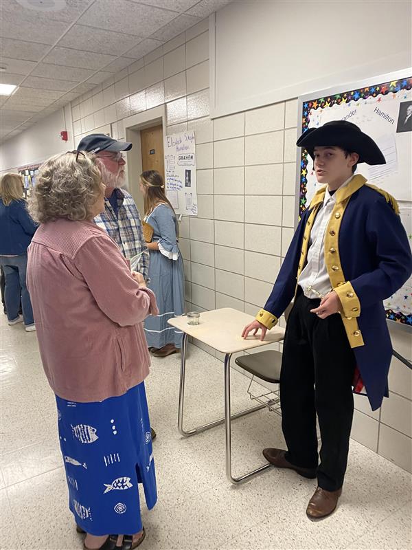 Adults stand in front of a student dressed in an old-fashioned jacket and hat with a poster behind them that reads "Alexander Hamilton."
