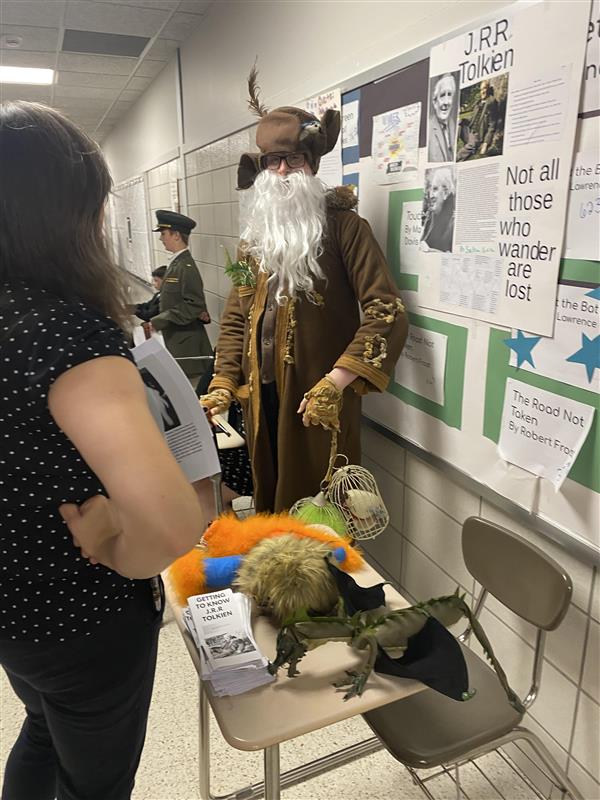 An adult stands in a hallway looking at a person dressed up in a brown robe with a large hat and long white beard. Behind the person is a poster that reads "J.R.R. Tolkien."
