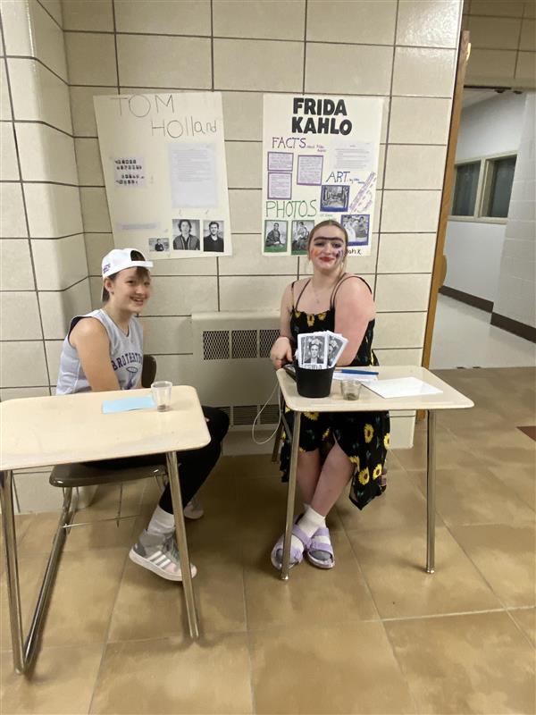 Two teenage students in costume sit at desks in a school hallway with posters behind them that say "Tom Holland" and "Frida Kahlo."