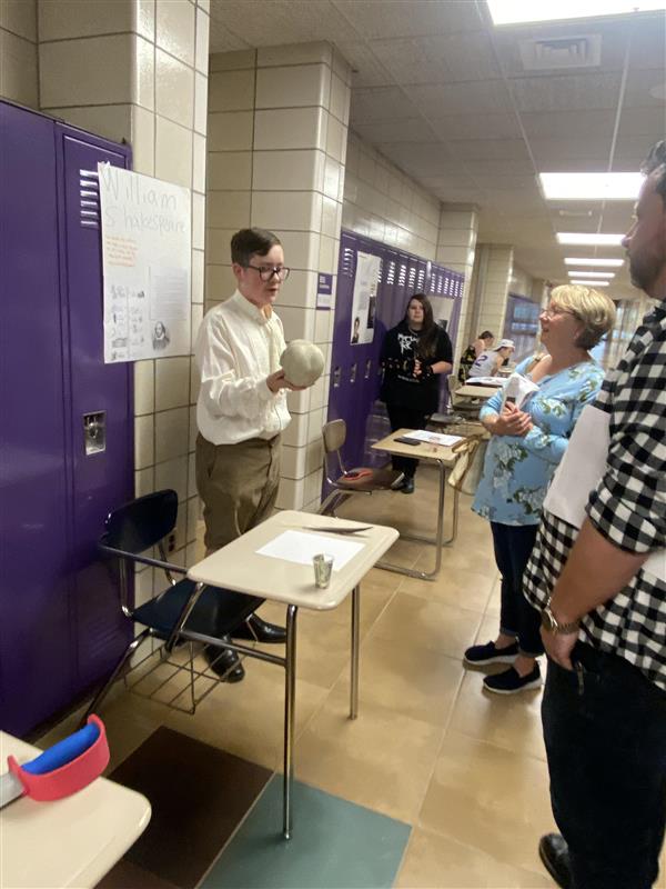 A student with glasses stands in front of a poster with the text "William Shakespeare" holding a skull in his right hand in a school hallway. 