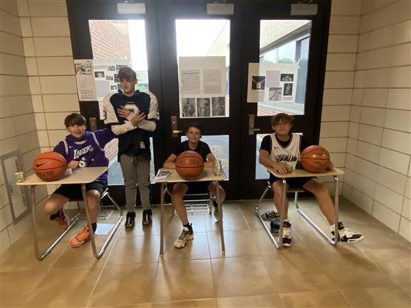 Preteen students wear basketball uniforms and sit at school desks in a hallway with basketballs. 