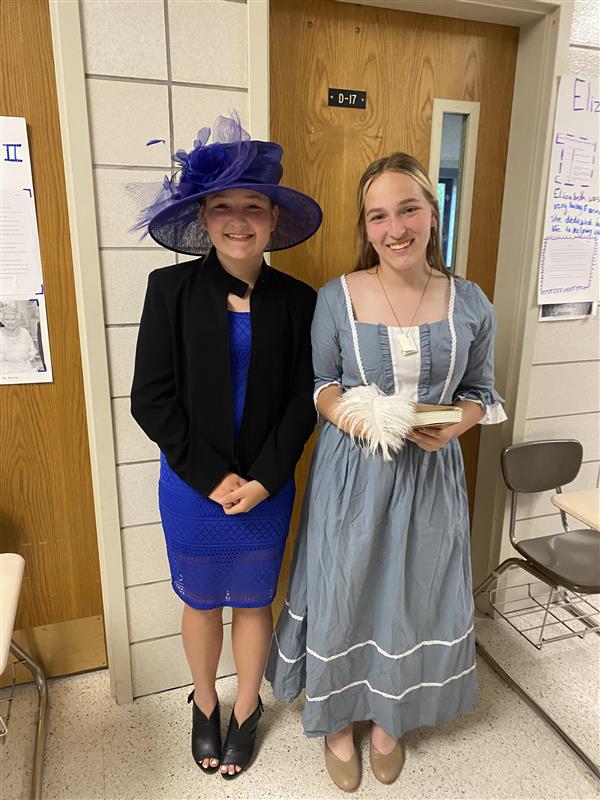 Two teenagers stand together in a school hallway. On the left a student wears a big blue hat with tulle, a black blazer, and a bright blue dress. On the right, a student wears a blue pilgrim-like dress and holds a feather pen.
