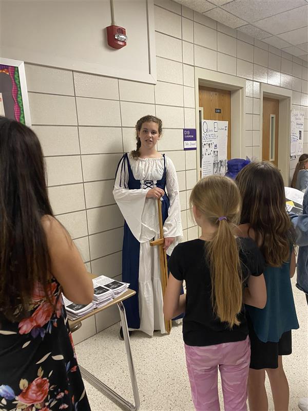 Elementary age students stand in a school hallway watching an older student in clothing from the middle ages holding a wooden cross.